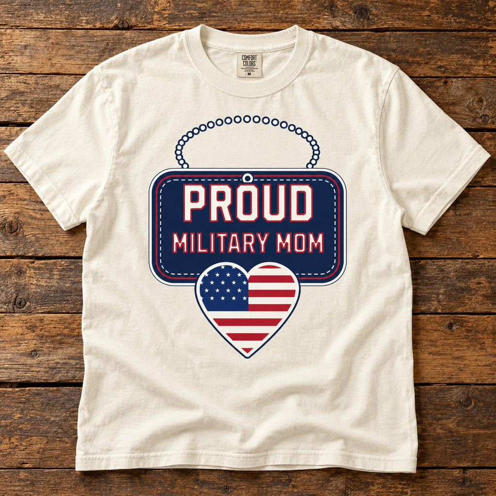 Proud Military Mom shirt with a navy dog tag style sign reading PROUD MILITARY MOM and an American flag heart, on an ivory tee laid on wood