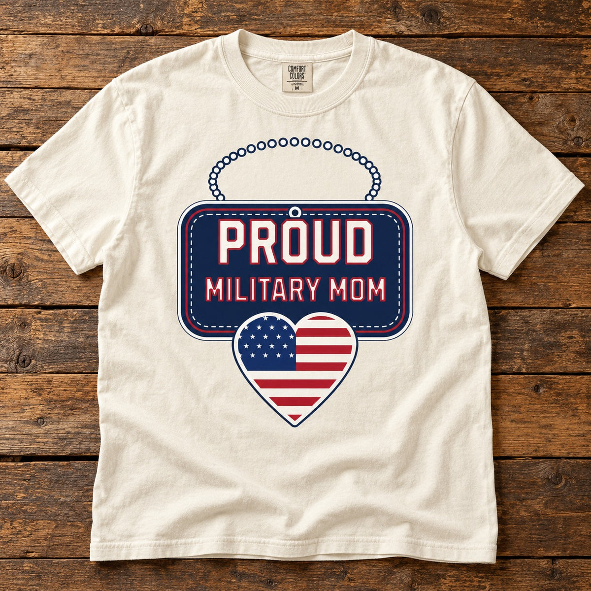 Proud Military Mom shirt with a navy dog tag style sign reading PROUD MILITARY MOM and an American flag heart, on an ivory tee laid on wood