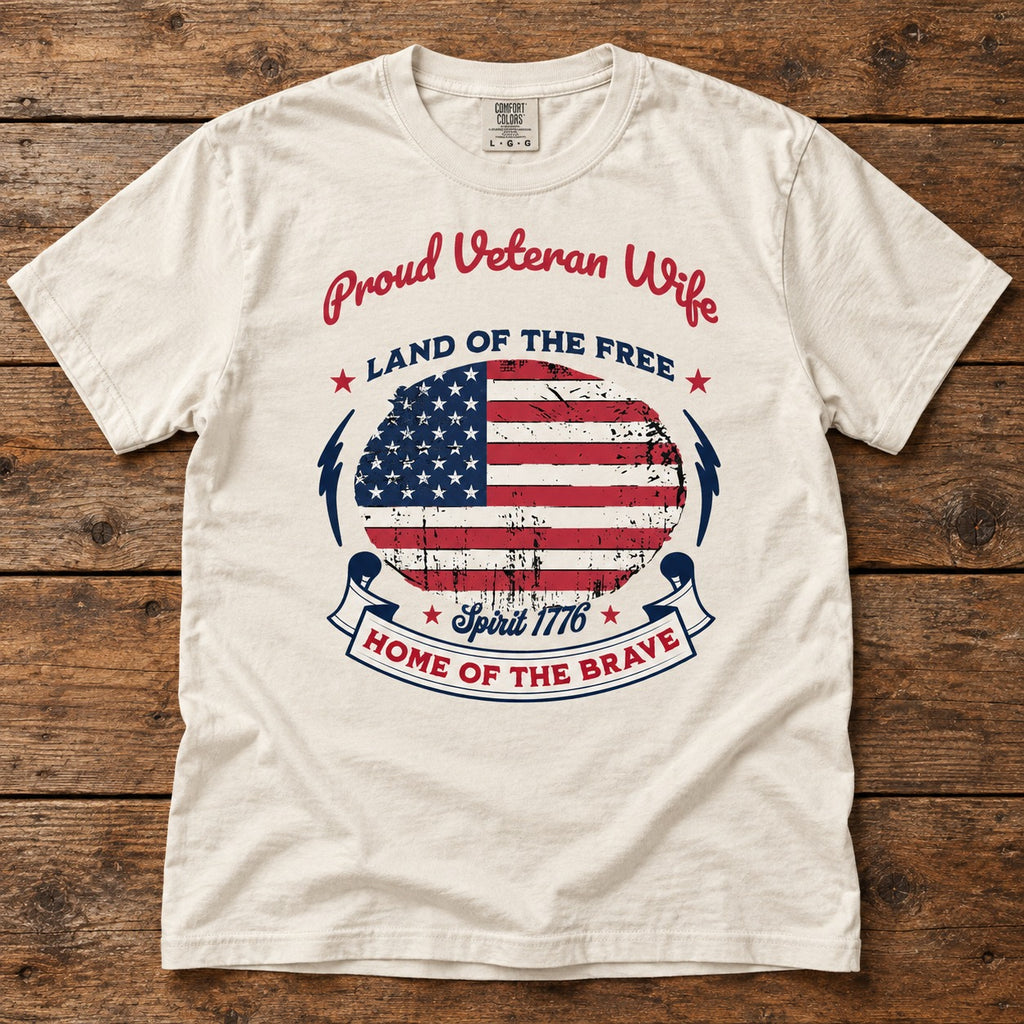 Proud Veteran Wife shirt with a distressed American flag circle, LAND OF THE FREE, Spirit 1776 and HOME OF THE BRAVE banner, on an ivory tee laid on wood